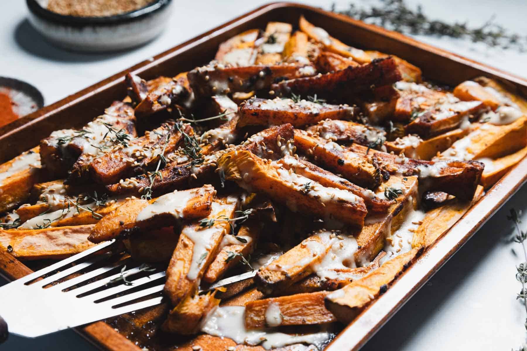 A baking sheet filled with thick-cut Maple Tahini Sweet Potatoes topped with creamy white sauce, sprigs of herbs, and ground pepper. A metal spatula rests under some of the fries.