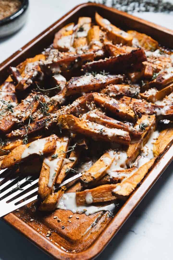 A baking tray filled with Maple Tahini Sweet Potatoes fries, topped with a drizzle of creamy white sauce, sprinkled with seeds and fresh herbs; a metal spatula rests beside the fries.
