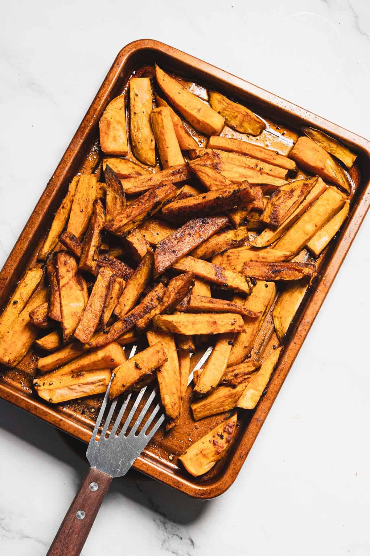A baking sheet with seasoned, roasted Maple Tahini Sweet Potatoes fries and a metal spatula on a white marble surface.