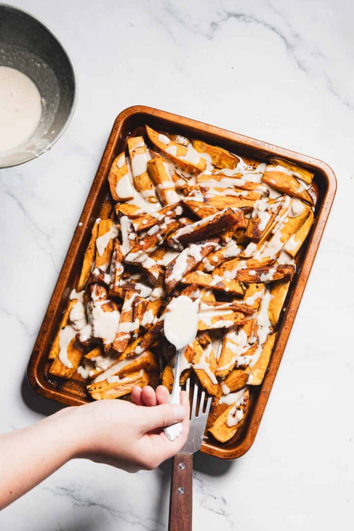A hand holds a fork over a baking tray of Maple Tahini Sweet Potatoes—seasoned wedges topped with creamy white sauce—set on a white marble surface. A bowl of extra sauce sits nearby.