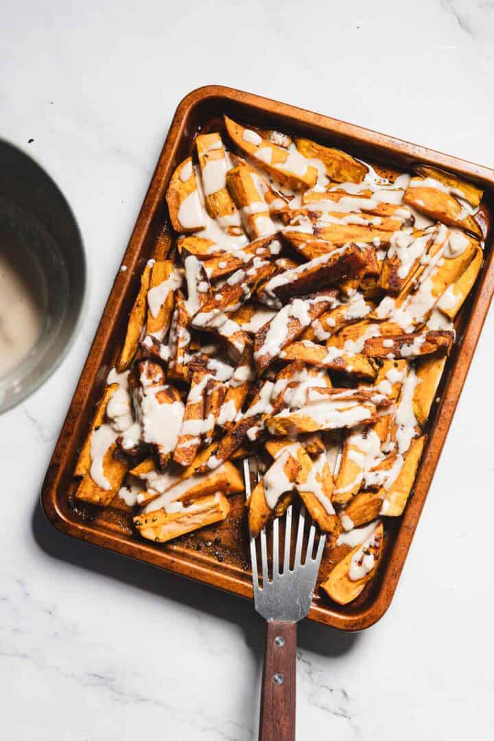 A baking tray filled with Maple Tahini Sweet Potatoes, drizzled with a creamy white sauce, rests on a white marble surface. A metal spatula sits in the tray, while a bowl is partially visible in the top left corner.