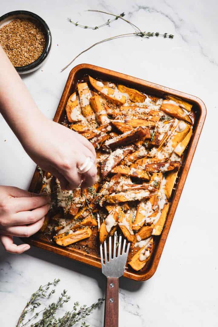 A person sprinkles herbs over a tray of Maple Tahini Sweet Potatoes topped with creamy sauce; a fork rests on the tray, with a small bowl of seasoning and fresh thyme on a marble surface nearby.