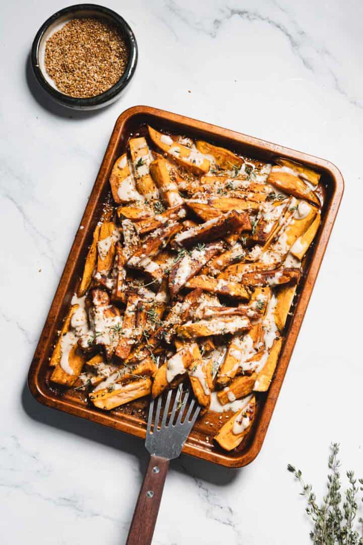 A baking sheet with golden-brown Maple Tahini Sweet Potatoes drizzled with creamy sauce and sprinkled with herbs. A metal spatula rests on the sheet, and a small bowl of seasoning sits nearby on a marble surface.