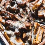 A close-up of Maple Tahini Sweet Potatoes on a tray—baked sweet potato fries topped with creamy white sauce, sprinkled with sesame seeds, and garnished with fresh herb sprigs.