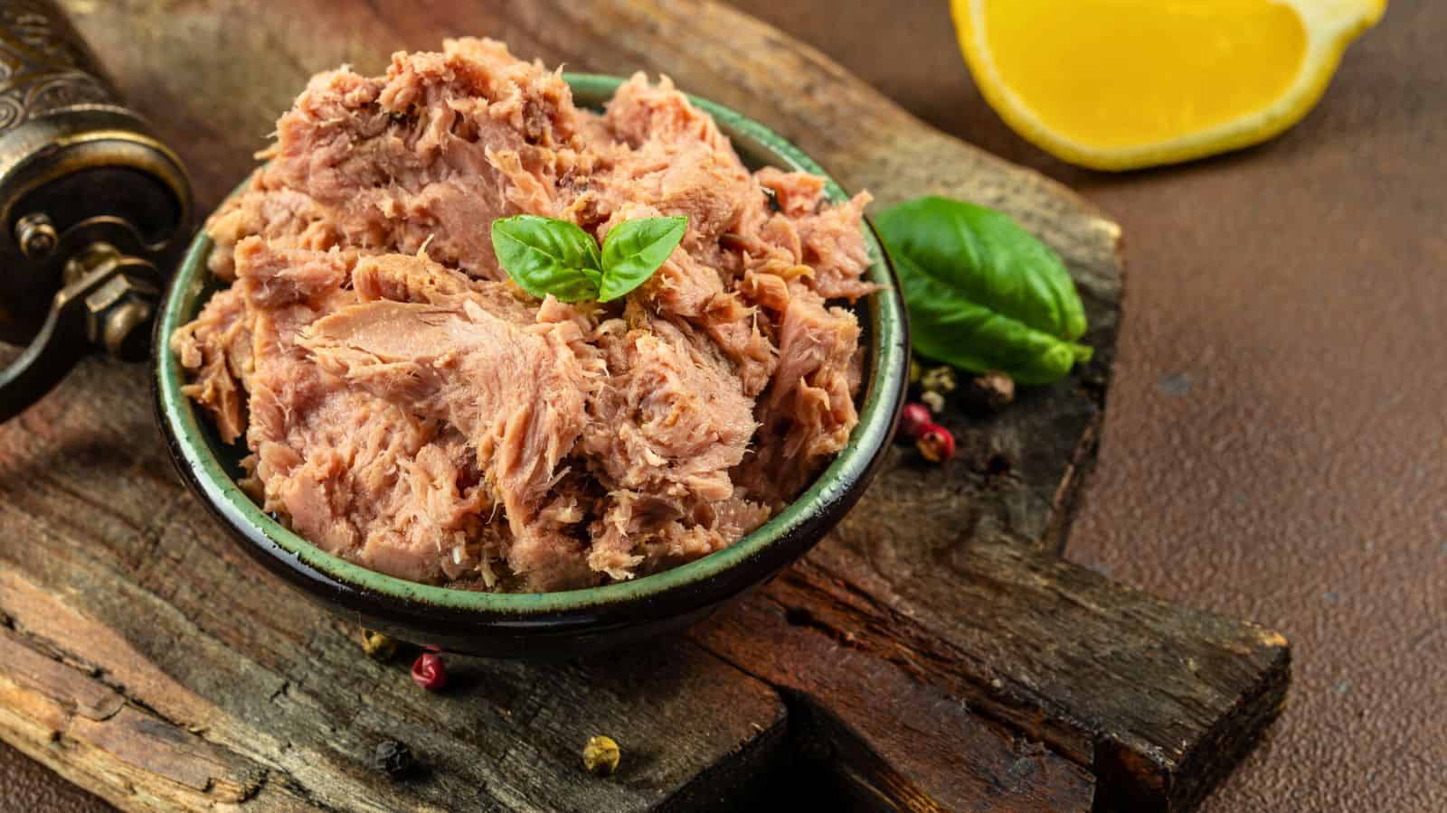 A bowl of flaked tuna garnished with a basil leaf sits on a rustic wooden board, with a lemon wedge and additional basil leaves in the background.