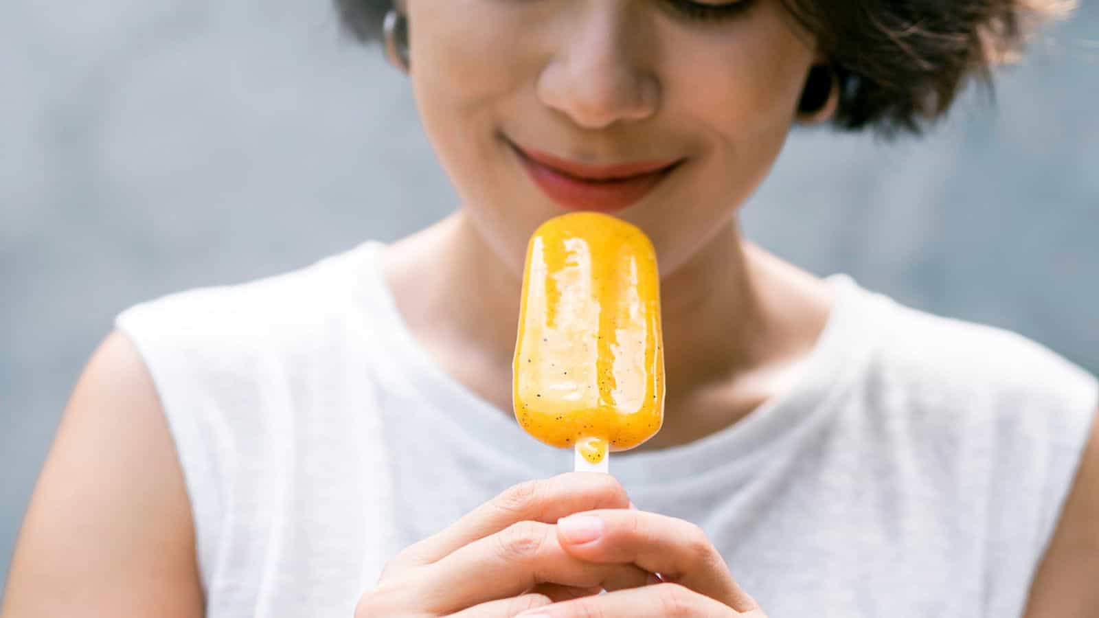 A person wearing a white sleeveless top holds a yellow popsicle close to their mouth and smiles slightly. The background is out of focus.