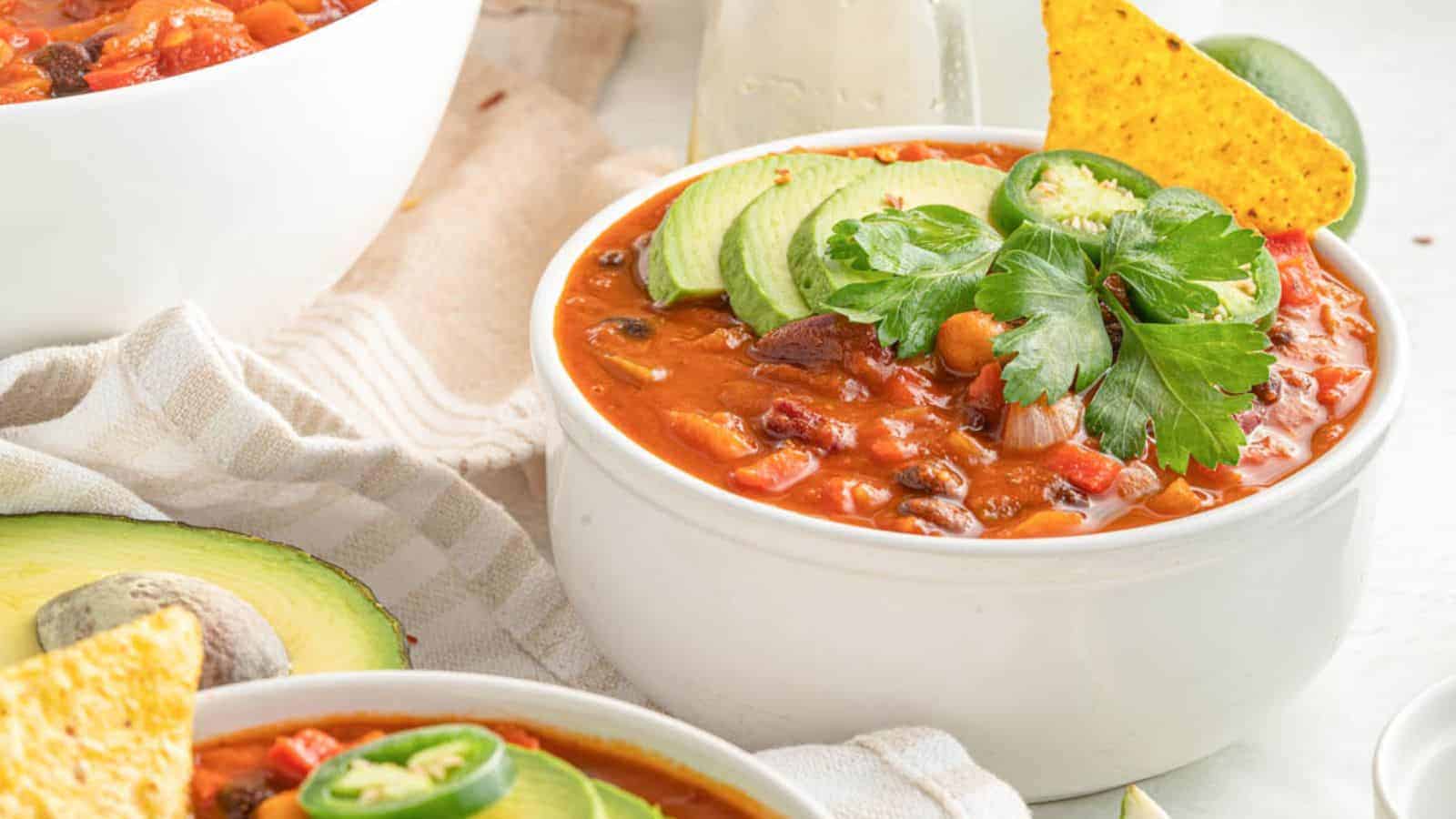 A white bowl of chili topped with avocado slices, jalape&ntilde;o, cilantro, and a tortilla chip sits on a light striped towel. Another bowl and avocado are partially visible nearby.