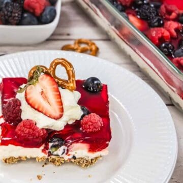 A dessert square with a pretzel crust, creamy filling, red gelatin topping, berries, whipped cream, and a pretzel rests on a white plate. A glass dish with more dessert and a bowl of berries are in the background.