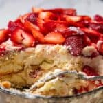 A close-up of a No-Bake Strawberry Tiramisu shows layers of cream, sponge cake, and sliced strawberries on top. A serving has been scooped out, revealing the dessert's delicious texture inside a glass dish.