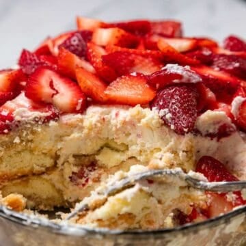 A close-up of a No-Bake Strawberry Tiramisu shows layers of cream, sponge cake, and sliced strawberries on top. A serving has been scooped out, revealing the dessert's delicious texture inside a glass dish.