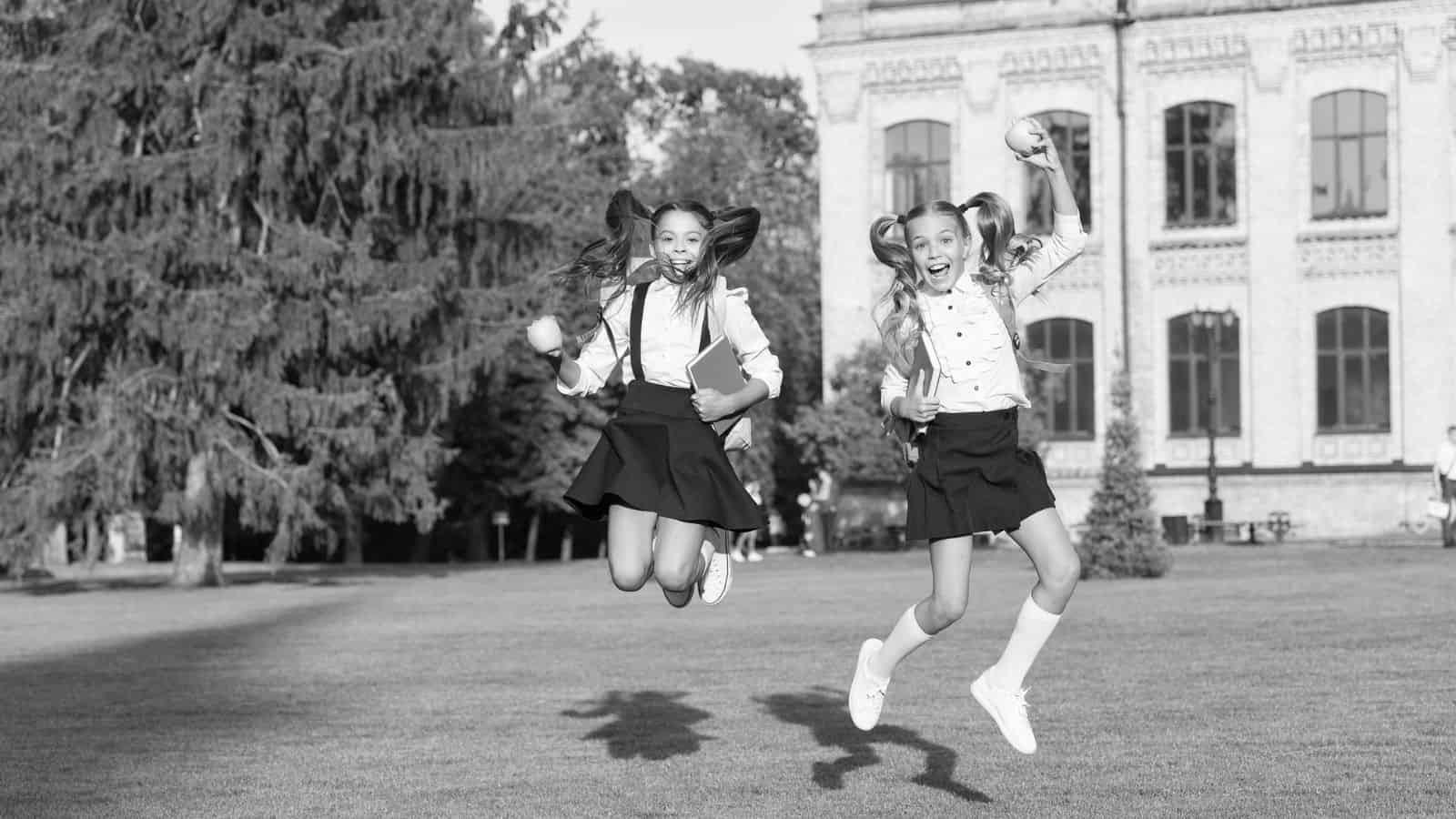 Two schoolgirls with pigtails, wearing uniforms and backpacks, are jumping in the air and smiling on a grassy area in front of a large building with tall windows. The photo is in black and white.