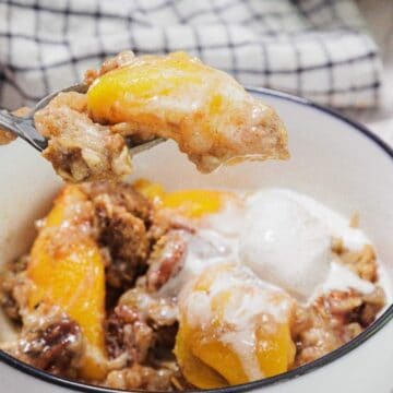 A bowl of peach cobbler topped with melting vanilla ice cream sits on a table. A spoon lifts a portion of the dessert, with chunks of peach and cobbler visible. A checkered cloth is in the background.