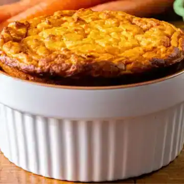 A golden-brown soufflé in a white ramekin sits on a wooden surface, surrounded by a spoon, fresh carrots, eggs in a carton, and a bit of eggshell.