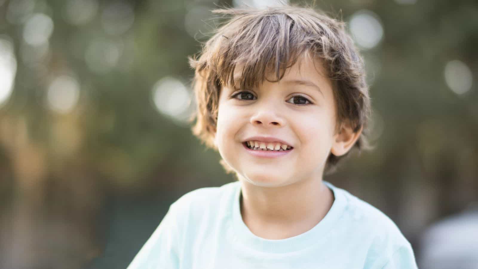 A young child with light brown hair and a light blue shirt smiles at the camera outdoors, with a blurred green background.