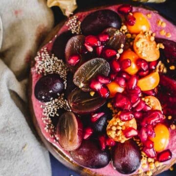A smoothie bowl topped with halved grapes, sliced plums, pomegranate seeds, golden berries, chia seeds, and bee pollen, placed next to a vintage spoon on a beige cloth.