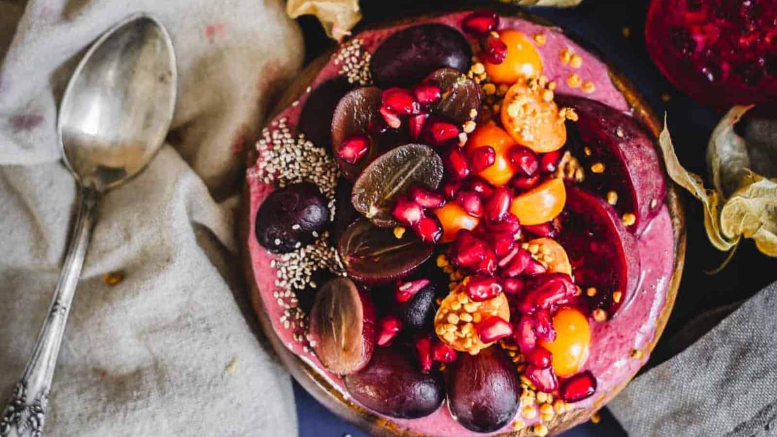 A smoothie bowl topped with halved grapes, sliced plums, pomegranate seeds, golden berries, chia seeds, and bee pollen, placed next to a vintage spoon on a beige cloth.