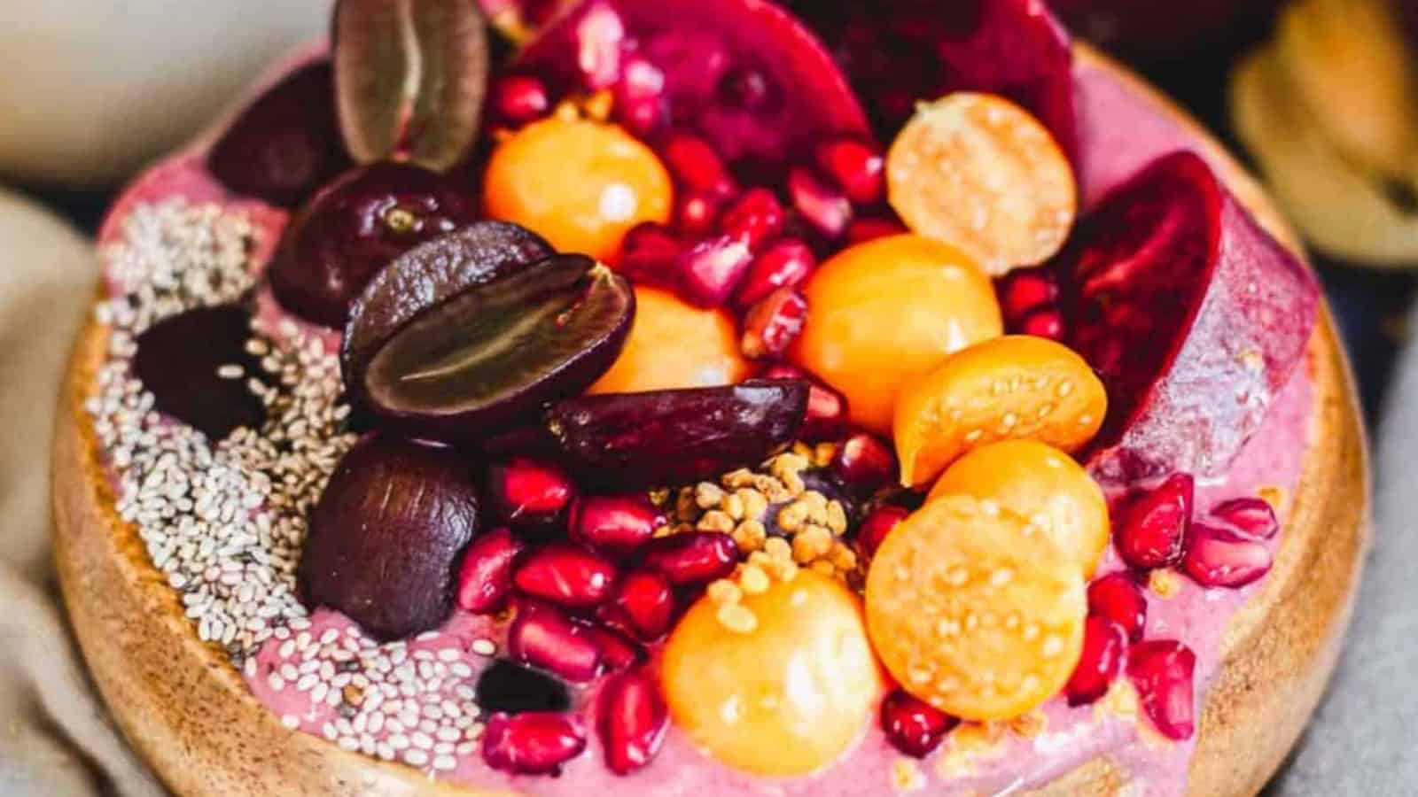 A smoothie bowl topped with sliced grapes, golden berries, pomegranate seeds, beet slices, chia seeds, and bee pollen, arranged in a wooden bowl.