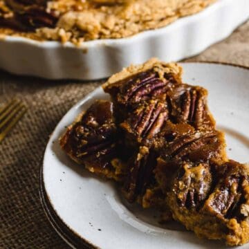 A white plate with two pieces of pecan pie sits on a fabric surface. Behind it is a partially visible pie in a white ceramic dish. A gold fork lies to the left of the plate.