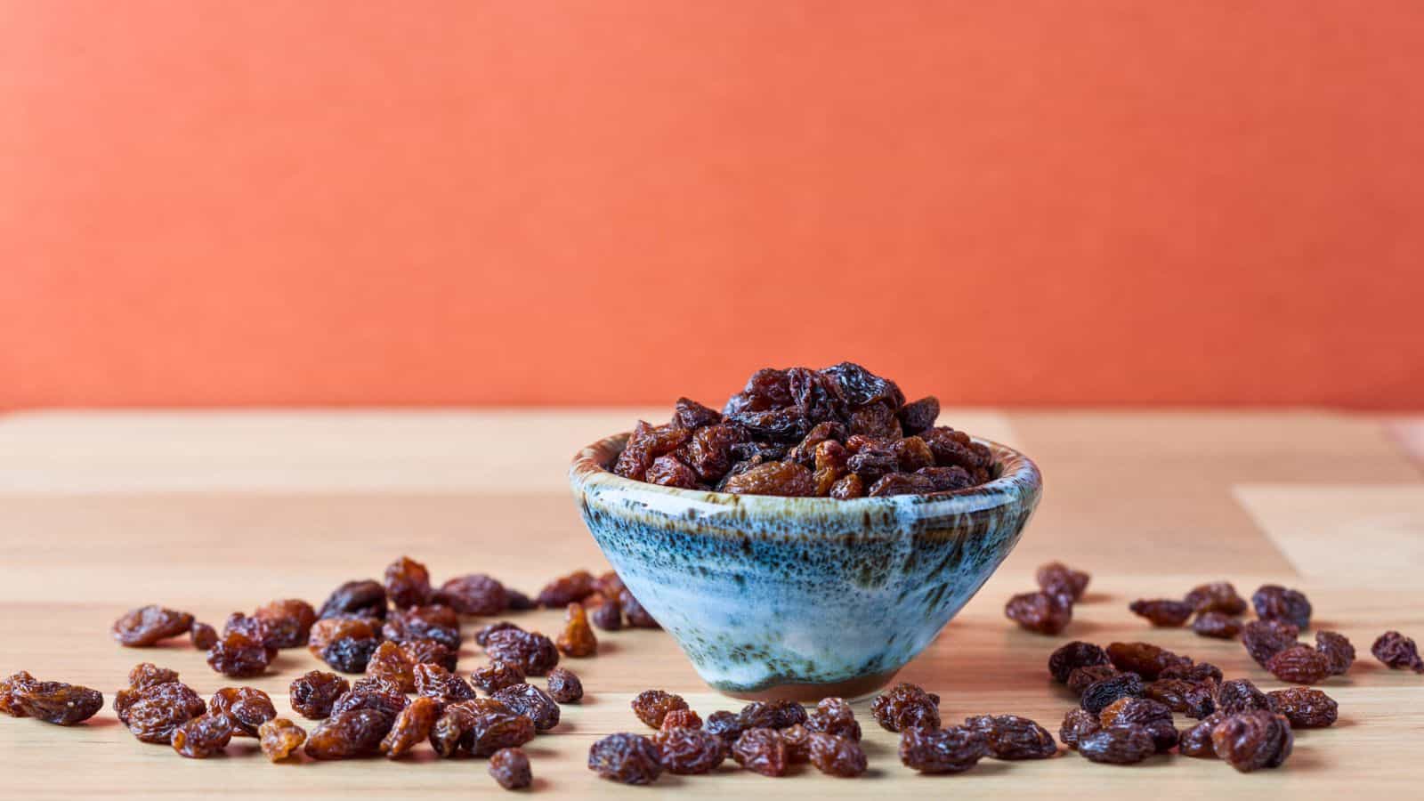 A small ceramic bowl filled with raisins sits on a wooden surface, with more raisins scattered around it. The background is an orange wall.