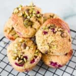 A stack of Raspberry Polenta Muffins with visible chunks of pistachios and red berry pieces sits on a black wire cooling rack against a light background.