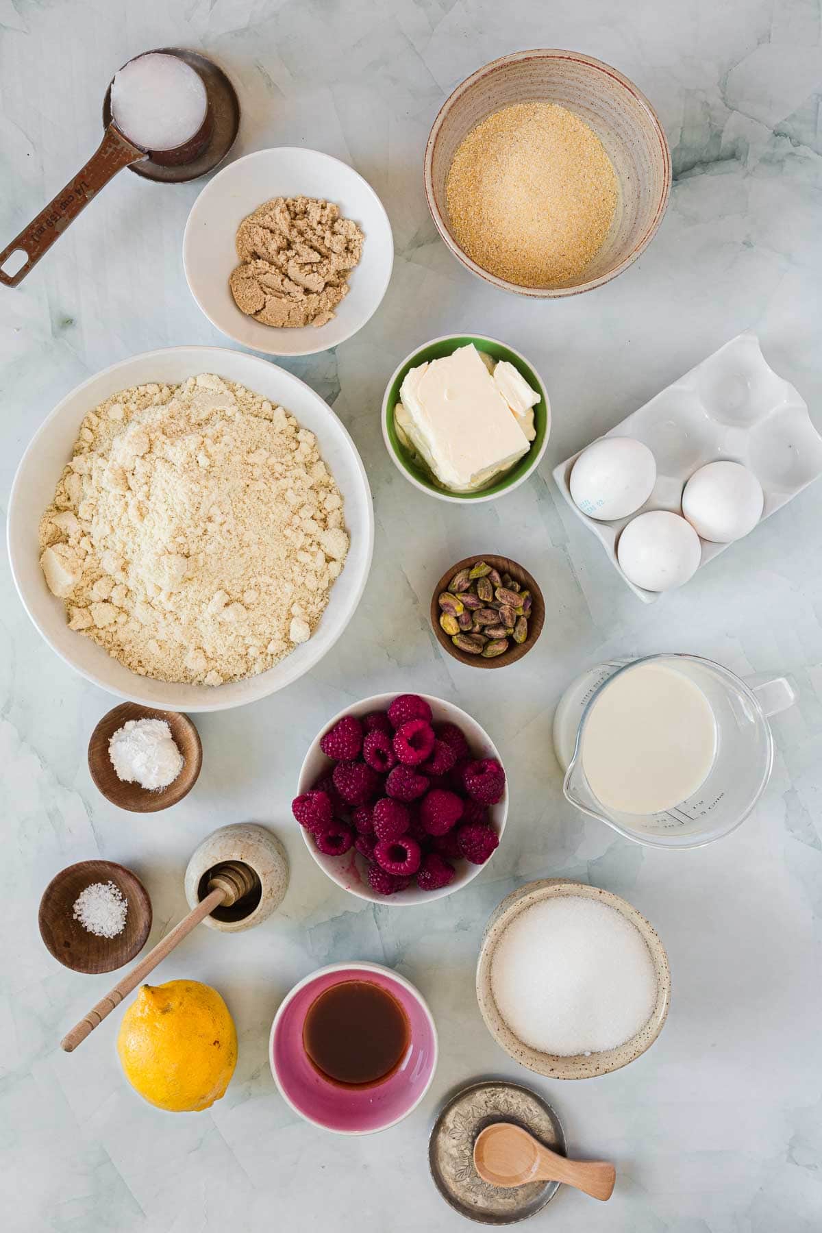 Assorted baking ingredients arranged on a light surface, perfect for making Raspberry Polenta Muffins—including flour, sugars, eggs, milk, butter, raspberries, a lemon, pistachios, vanilla extract, salt, and a spice jar with a spoon.