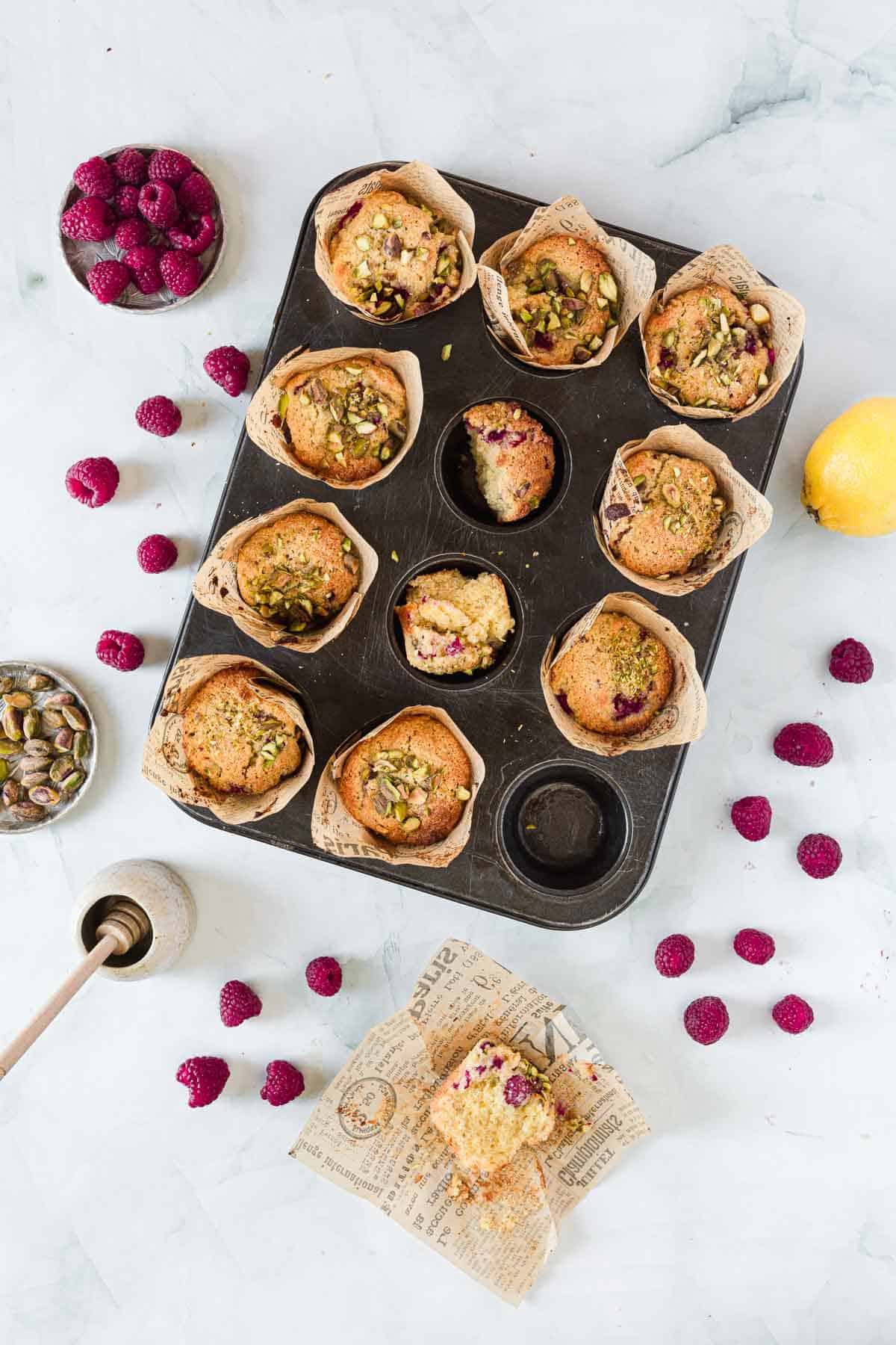 A muffin tin holds eight baked Raspberry Polenta Muffins with paper liners, one space is empty, and one muffin is partially eaten. Fresh raspberries, a lemon, pistachios, honey dipper, and a muffin on paper are arranged around the tin.