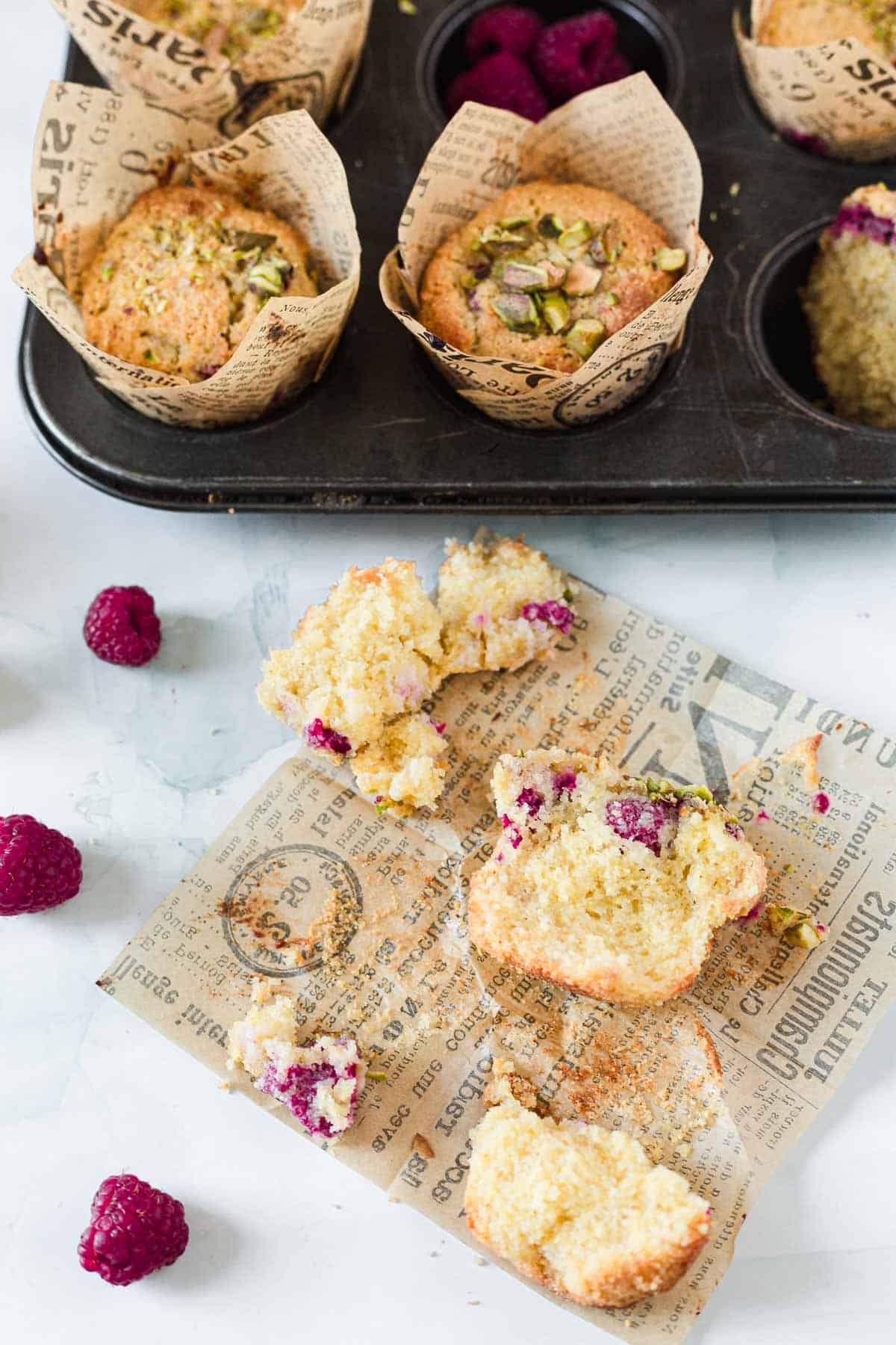 A broken Raspberry Polenta Muffin sits on printed parchment paper with whole raspberries nearby. In the background, a muffin tin holds more Raspberry Polenta Muffins topped with chopped pistachios.