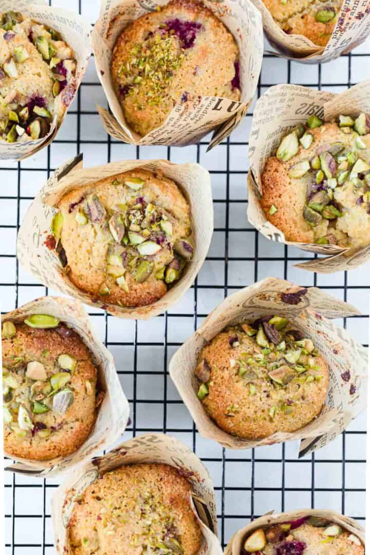 Seven Raspberry Polenta Muffins in paper wrappers, topped with chopped pistachios, are arranged on a cooling rack. Some muffins have visible berry pieces. The wrappers feature a newspaper print design.