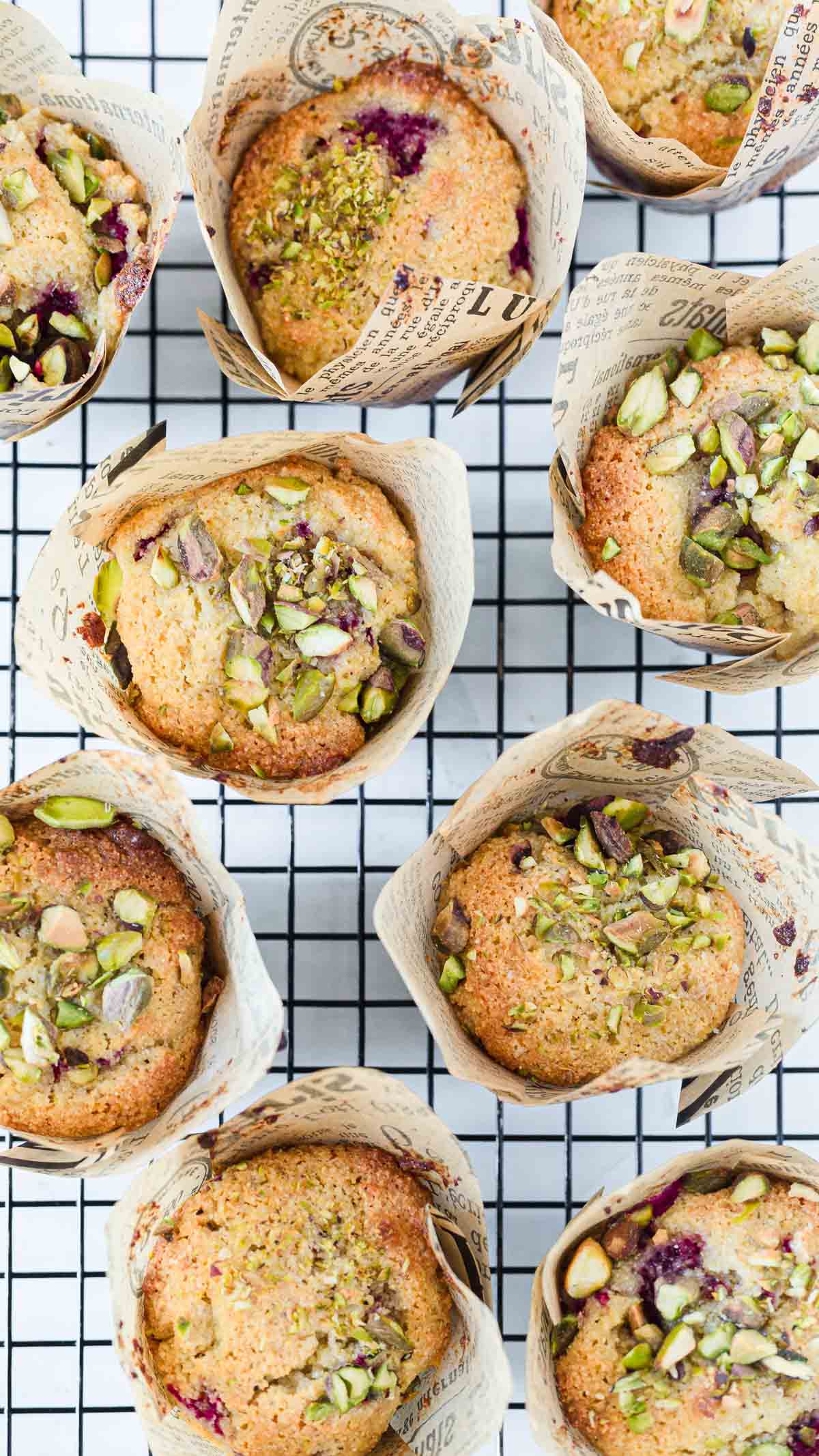 Nine Raspberry Polenta Muffins in decorative paper wrappers, topped with chopped pistachios, are arranged on a black wire cooling rack. The golden-brown muffins have a rustic look and reveal hints of berry filling.