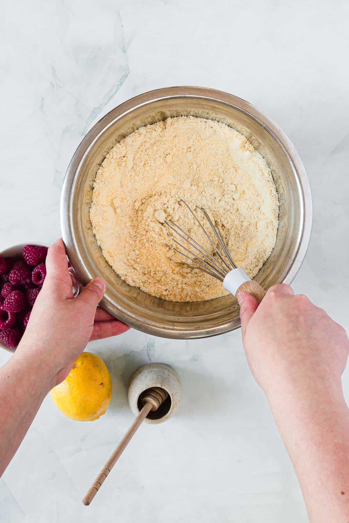 A person whisks dry ingredients in a metal mixing bowl, preparing for Raspberry Polenta Muffins. Nearby are a bowl of raspberries, a whole lemon, and a small container of honey with a dipper on a white countertop.