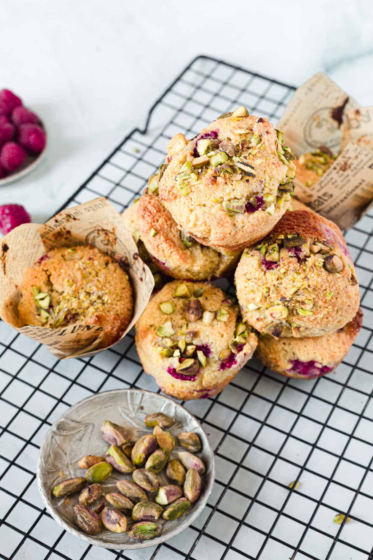 A stack of Raspberry Polenta Muffins topped with chopped pistachios and raspberries sits on a cooling rack, with a bowl of pistachios and a small dish of raspberries nearby. Some muffins are wrapped in parchment paper.