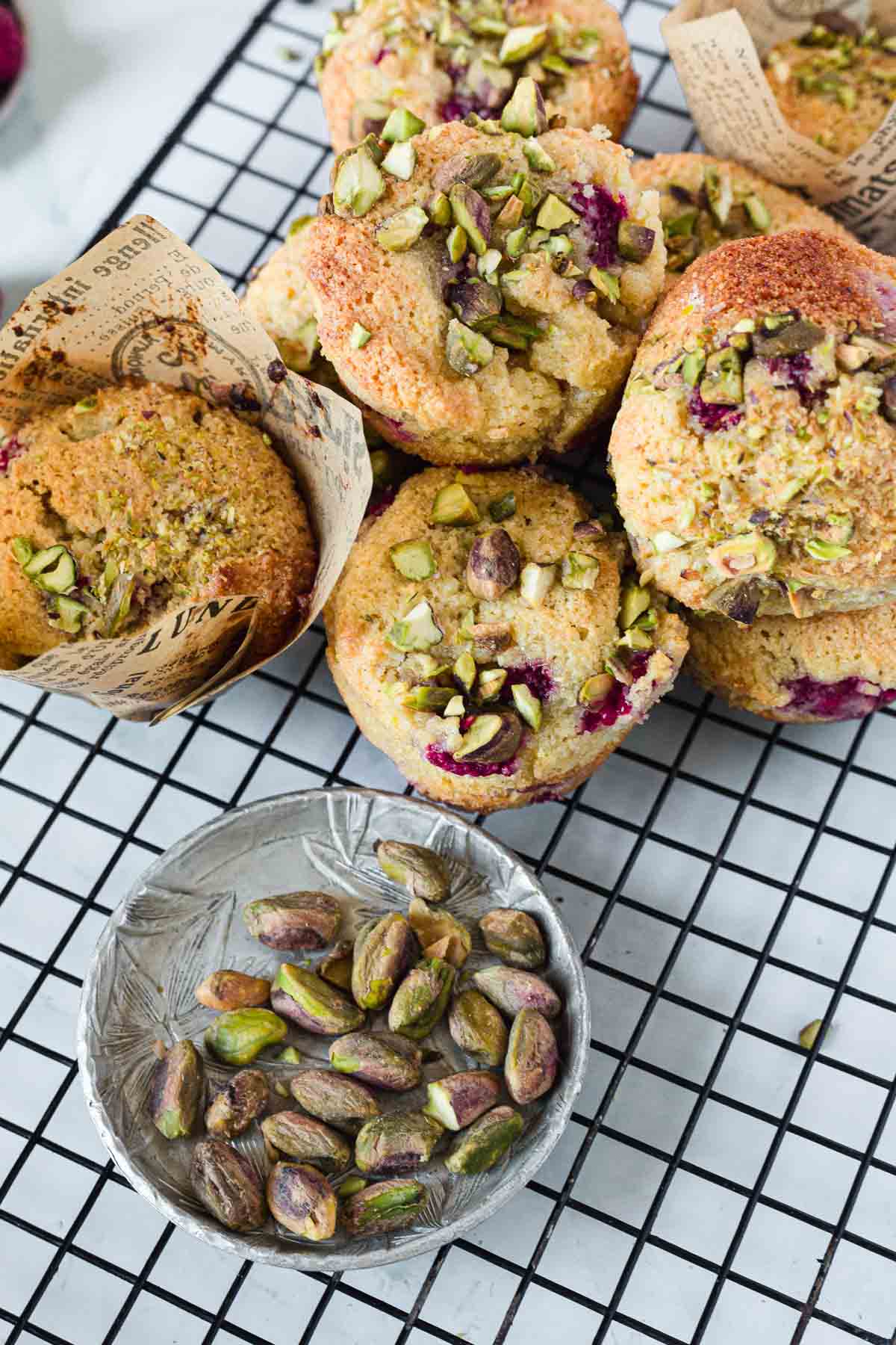 A group of Raspberry Polenta Muffins topped with chopped pistachios sits on a cooling rack, some in decorative paper liners. A small bowl filled with whole pistachios is placed nearby.