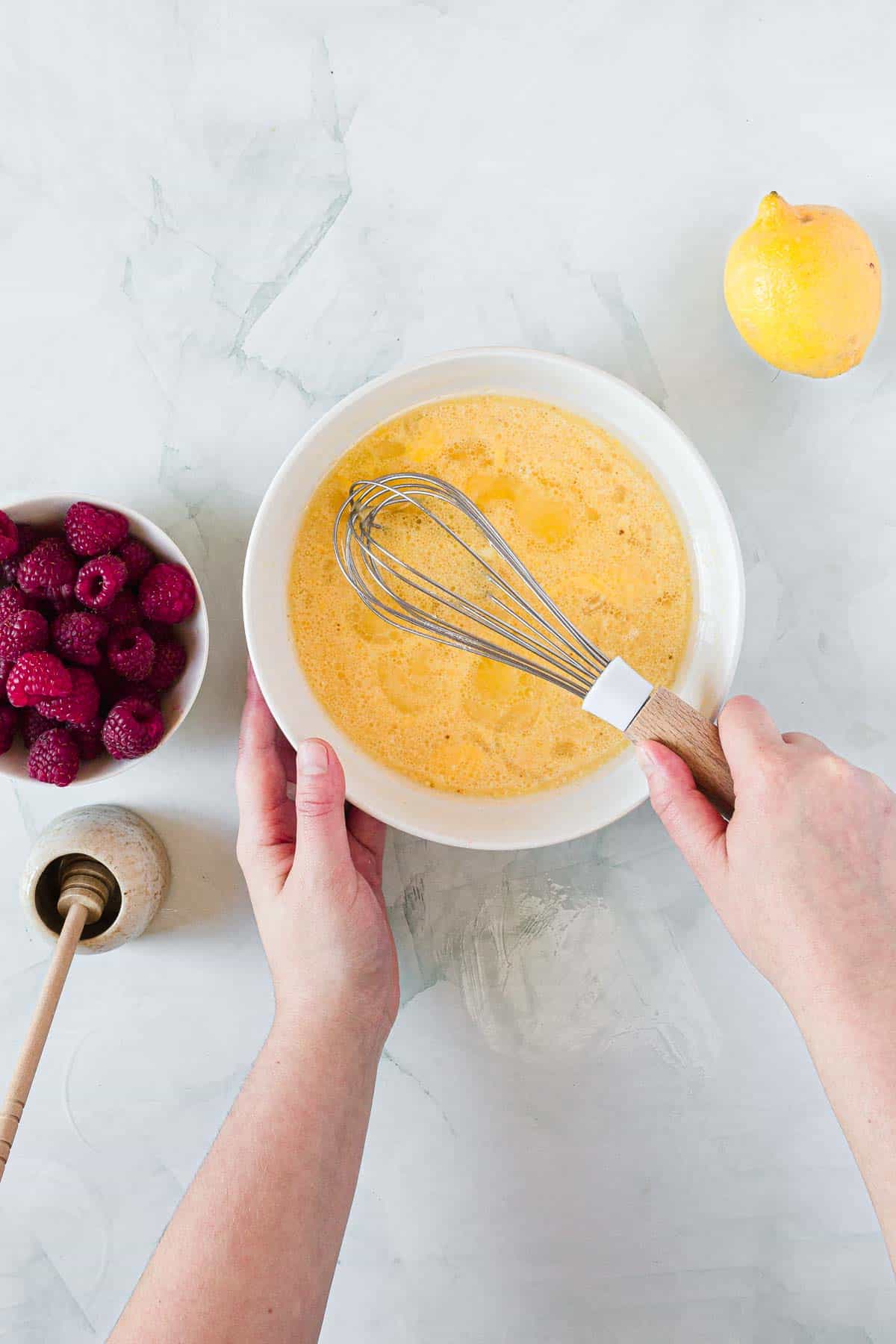 A person is whisking a yellow mixture in a white bowl, preparing batter for Raspberry Polenta Muffins. Nearby, there’s a bowl of raspberries, a honey dipper with honey, and a whole lemon on a light surface.