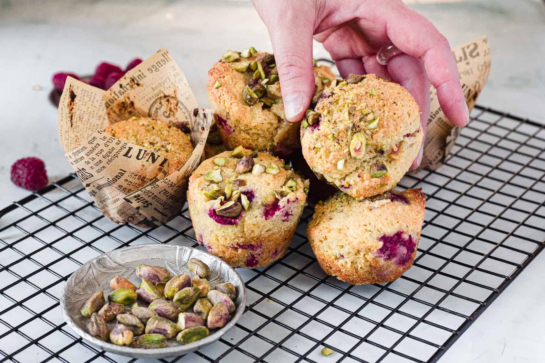 A hand picks up a Raspberry Polenta Muffin topped with chopped pistachios from a cooling rack. Some muffins are wrapped in decorative paper, and a small dish of shelled pistachios sits nearby.