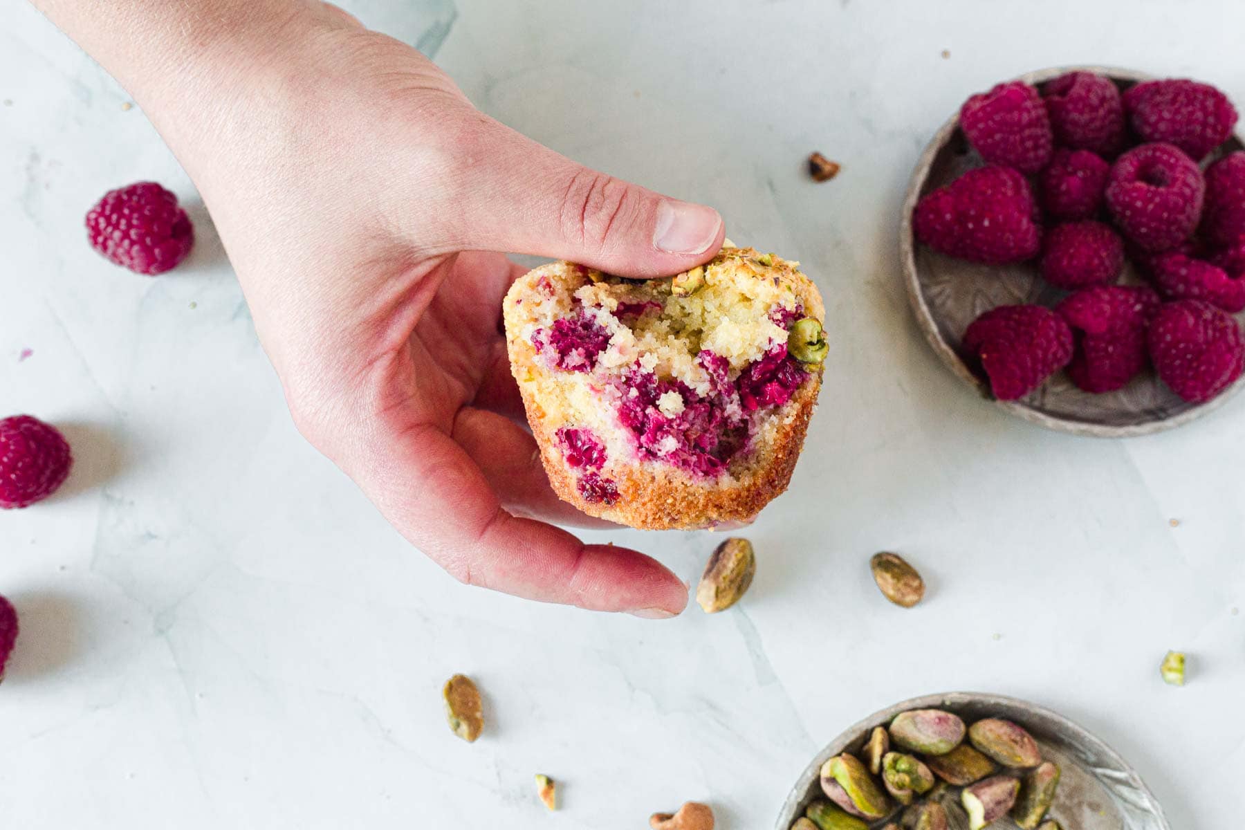 A hand holds a half-eaten Raspberry Polenta Muffin with visible raspberries and pistachios. Nearby, bowls of fresh raspberries and shelled pistachios rest on a white surface.