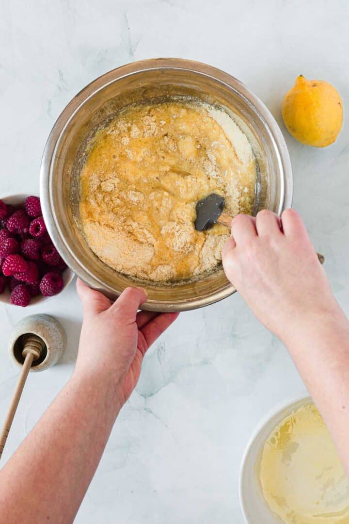 A person stirs a thick yellow batter for Raspberry Polenta Muffins in a metal bowl with a spatula. Nearby are a bowl of raspberries, a lemon, a honey dipper, and a white bowl with liquid on a light marble surface.