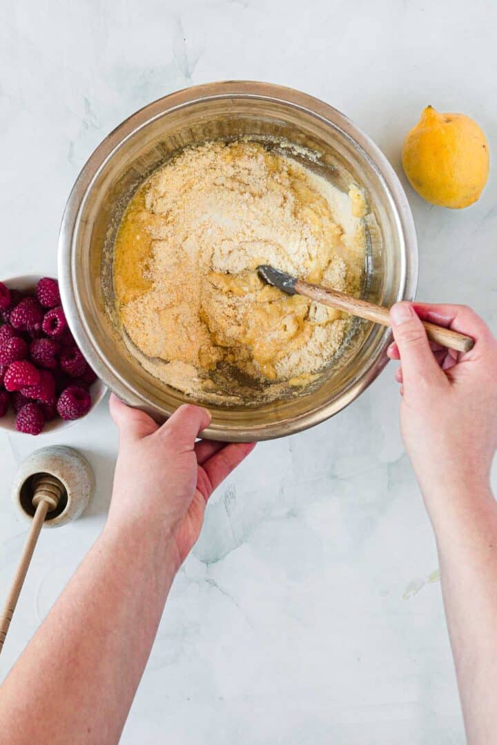 A pair of hands mixing batter for Raspberry Polenta Muffins with a wooden spoon in a metal bowl. Nearby are a lemon, a bowl of raspberries, and a honey dipper with honey on a white surface.