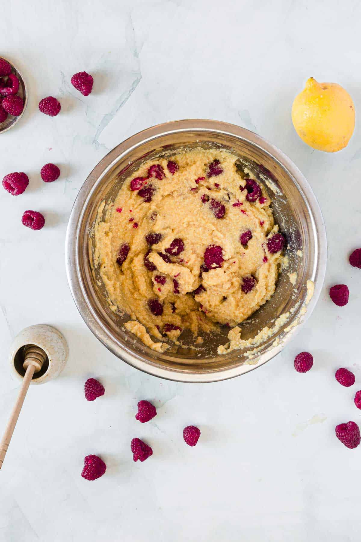 A metal mixing bowl holds yellow batter with raspberries mixed in, ready to become Raspberry Polenta Muffins. Loose raspberries, a lemon, and a honey dipper are arranged on the white countertop around the bowl.