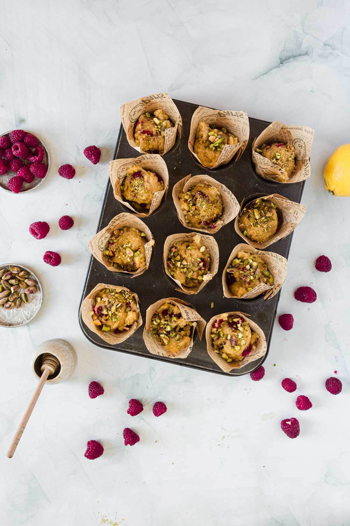 A muffin tin holding 12 Raspberry Polenta Muffins in brown paper wrappers, garnished with pistachios and raspberries. Surrounding the tin are scattered raspberries, a lemon, bowls of pistachios and raspberries, and a honey dipper.