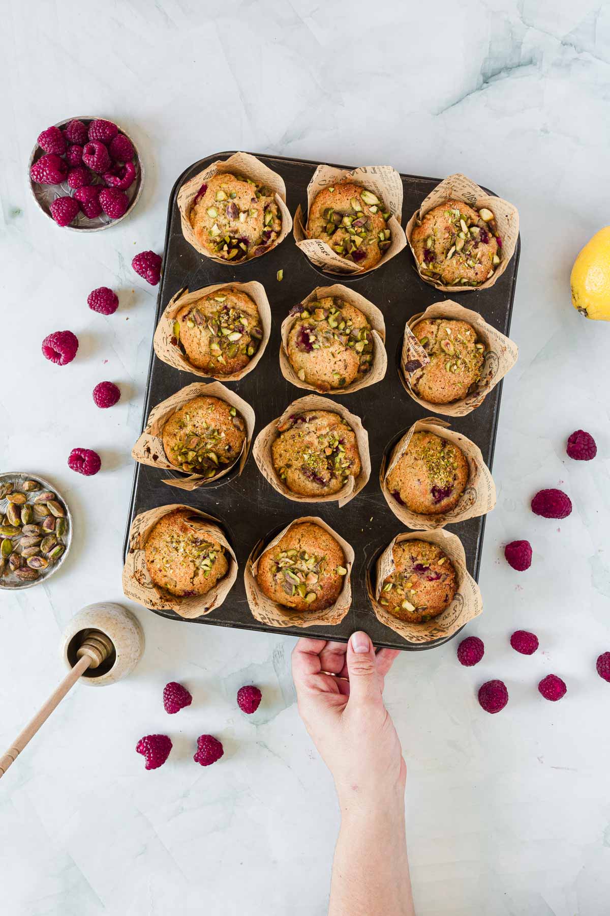 A hand holds a muffin tin with ten Raspberry Polenta Muffins, topped with pistachios and raspberries. Surrounding the tin are loose raspberries, a lemon, a bowl of pistachios, a small jug, and a bowl of raspberries on a white surface.