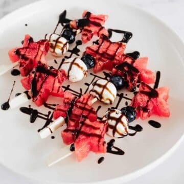 Watermelon and blueberry skewers with mozzarella balls are arranged on a white plate, drizzled with balsamic glaze. The skewers are placed on a marble surface.