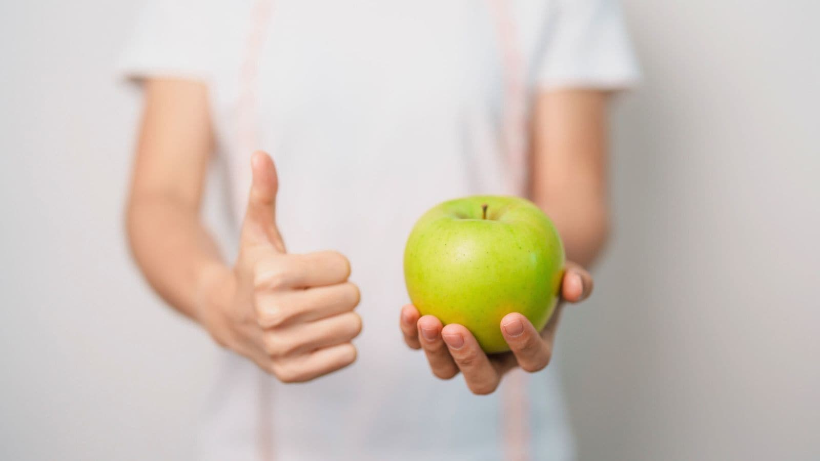 A person wearing a white shirt holds a green apple in one hand and gives a thumbs-up with the other hand. The background is plain and out of focus.