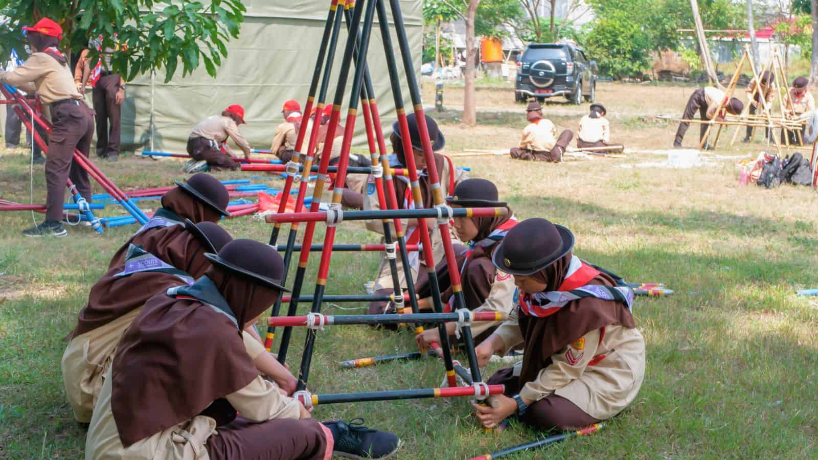 A group of scouts in brown uniforms and hats are assembling structures with rods on a grassy field. Some scouts are sitting in groups, while others work in the background near a tent and parked cars.