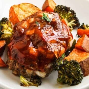 A white plate with a serving of meatloaf topped with sauce, surrounded by roasted broccoli, sweet potato chunks, and a halved roasted potato. A yellow napkin is partially visible on the side.