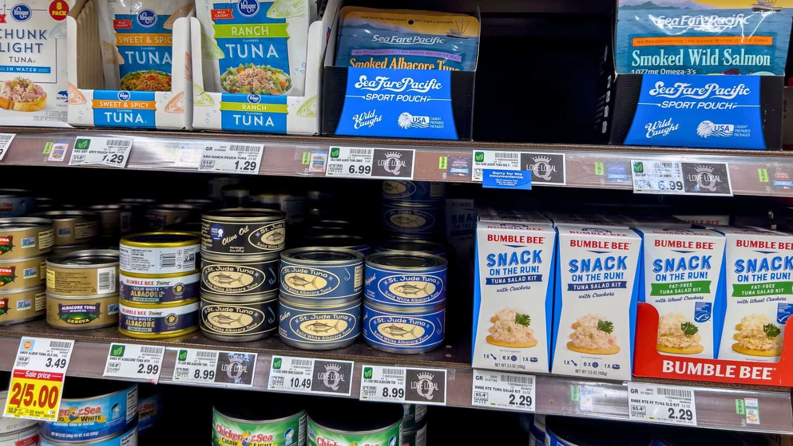 Grocery store shelves displaying various canned tuna and salmon products, including Bumble Bee Snack On The Run, Blue Harbor, and Sea Fare Pacific brands, with price tags visible below the items.