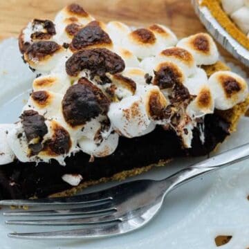 A slice of chocolate pie topped with toasted marshmallows sits on a white decorative plate with a fork. The remaining pie is partially visible in the background on a wooden surface.