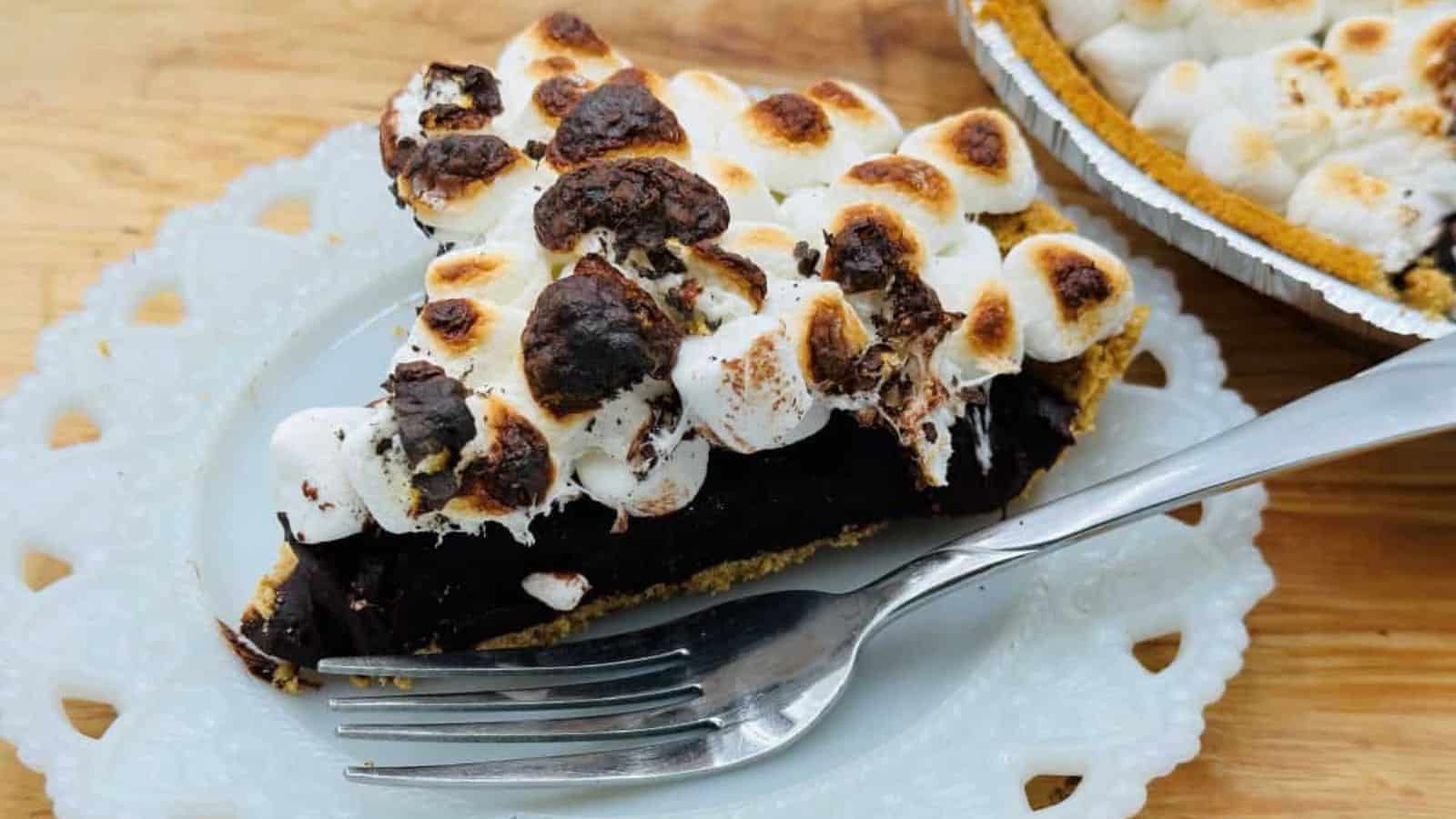 A slice of chocolate pie topped with toasted marshmallows on a white ornate plate, with a fork placed beside it, rests on a wooden surface. Another partially visible pie is in the background.