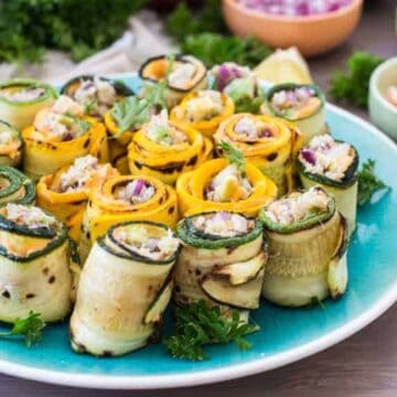 A blue plate holds rolled slices of yellow and green zucchini filled with a vegetable mixture, garnished with parsley—colorful pool party bites. Bowls of condiments and herbs are in the background on a light-colored table.