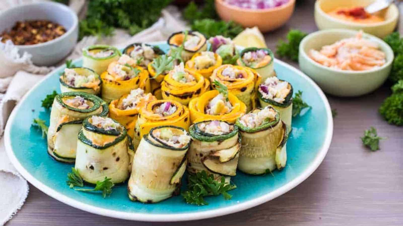 A blue plate holds rolled slices of yellow and green zucchini filled with a vegetable mixture, garnished with parsley—colorful pool party bites. Bowls of condiments and herbs are in the background on a light-colored table.