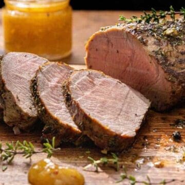 Sliced roasted pork tenderloin with herbs on a wooden cutting board, garnished with fresh thyme and black peppercorns, with a jar of yellow sauce in the background.